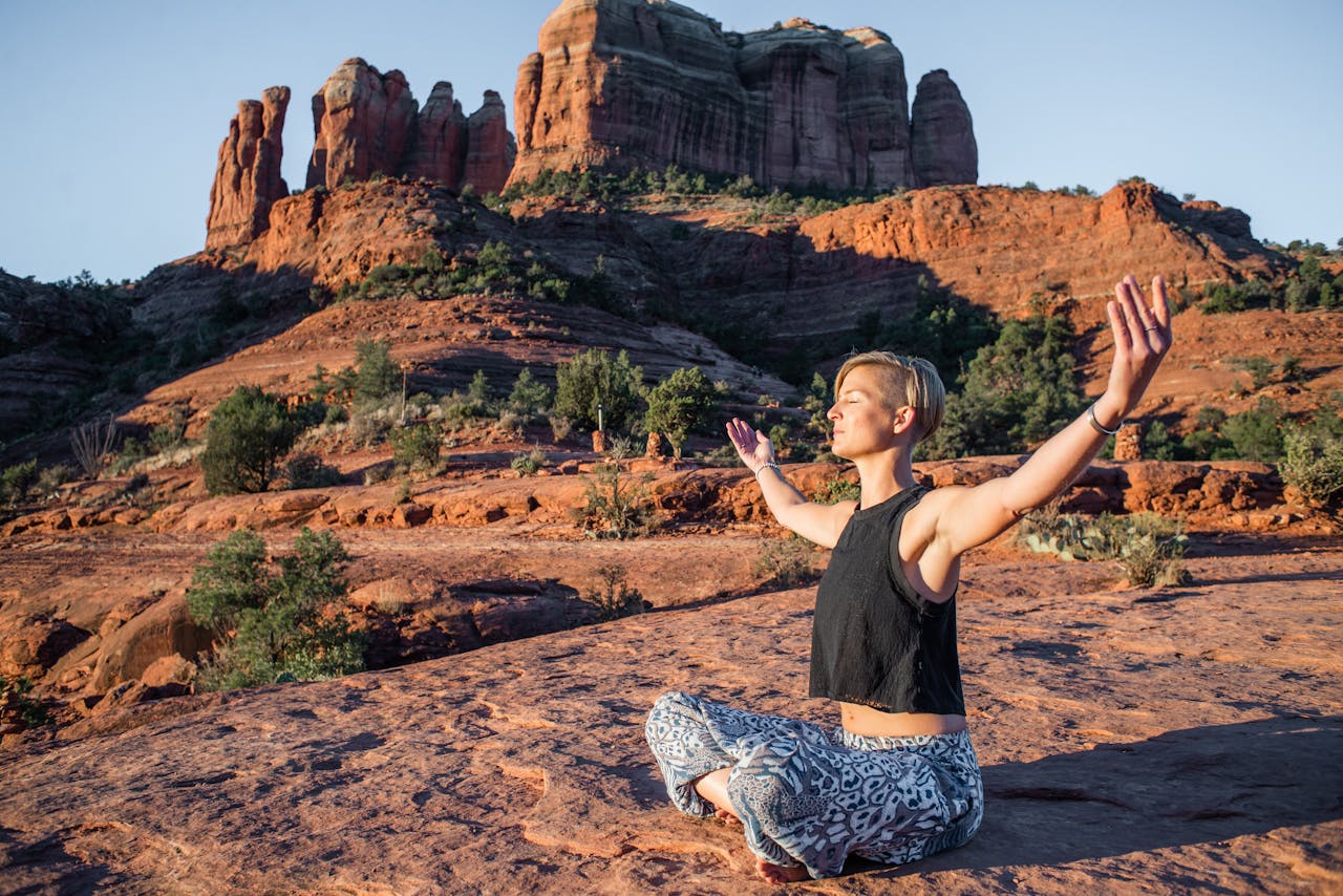 Young slim lady in summer top and hippie pants sitting with arms spread and eyes closed on rough ground while doing yoga near magnificent sunlit canyons